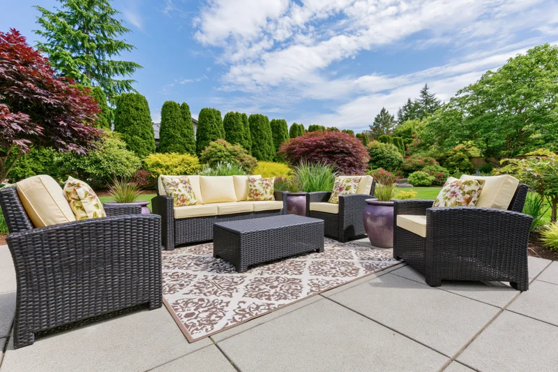 Dark wicker patio furniture set with beige cushions arranged on a patterned rug in a landscaped backyard garden