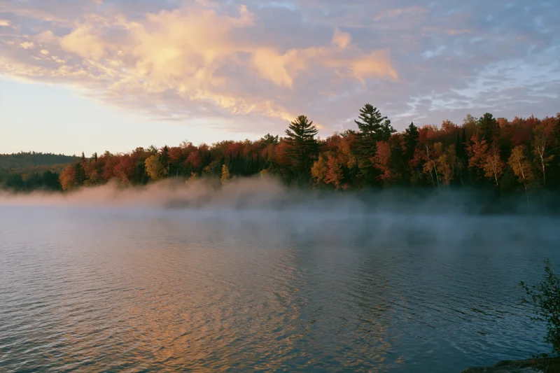 Calm lake reflecting autumn foliage under thin morning mist along forested shoreline