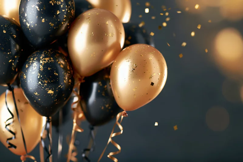 Cluster of gold and black metallic helium balloons with curling ribbons and falling confetti against a dark background with bokeh lights