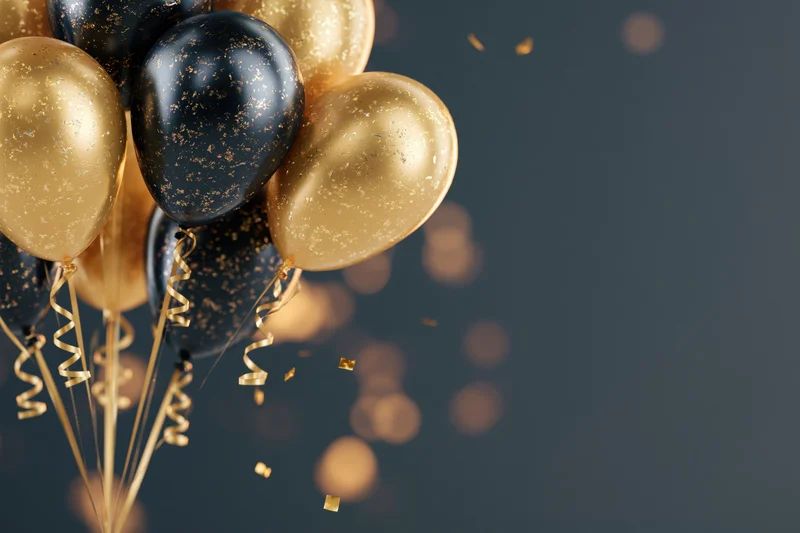 Cluster of gold and black metallic helium balloons with curling ribbons and falling confetti against a dark background with bokeh lights