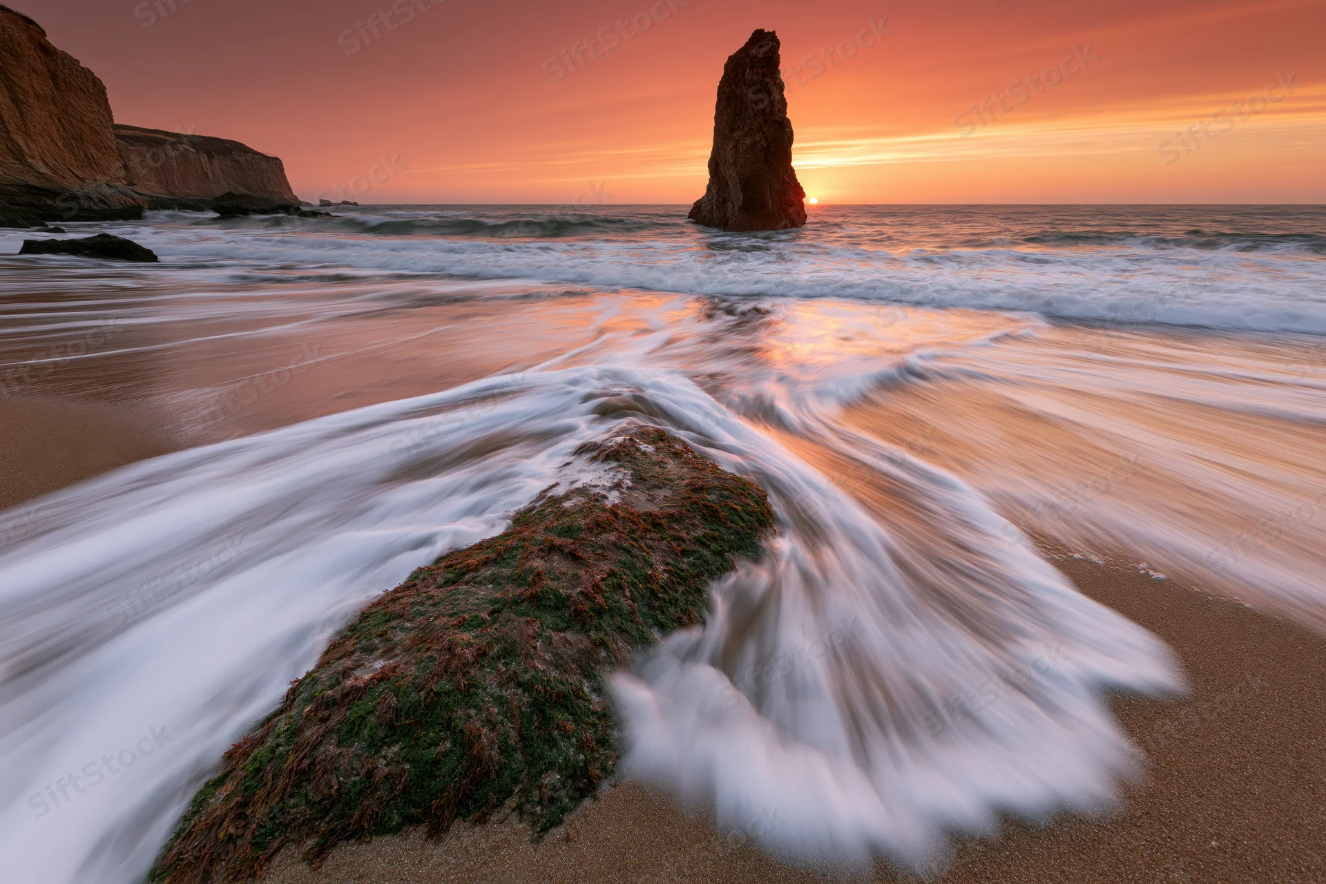 Ocean waves washing over moss-covered rock on sandy beach at sunrise