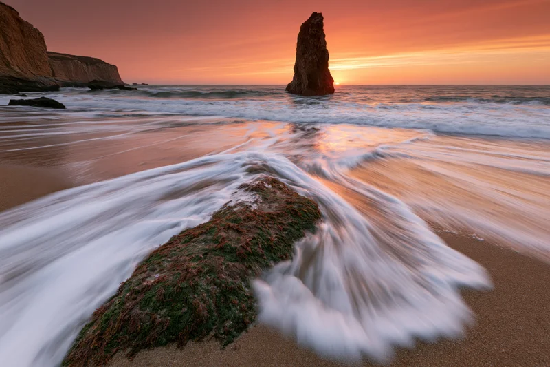 Ocean waves washing over moss-covered rock on sandy beach at sunrise
