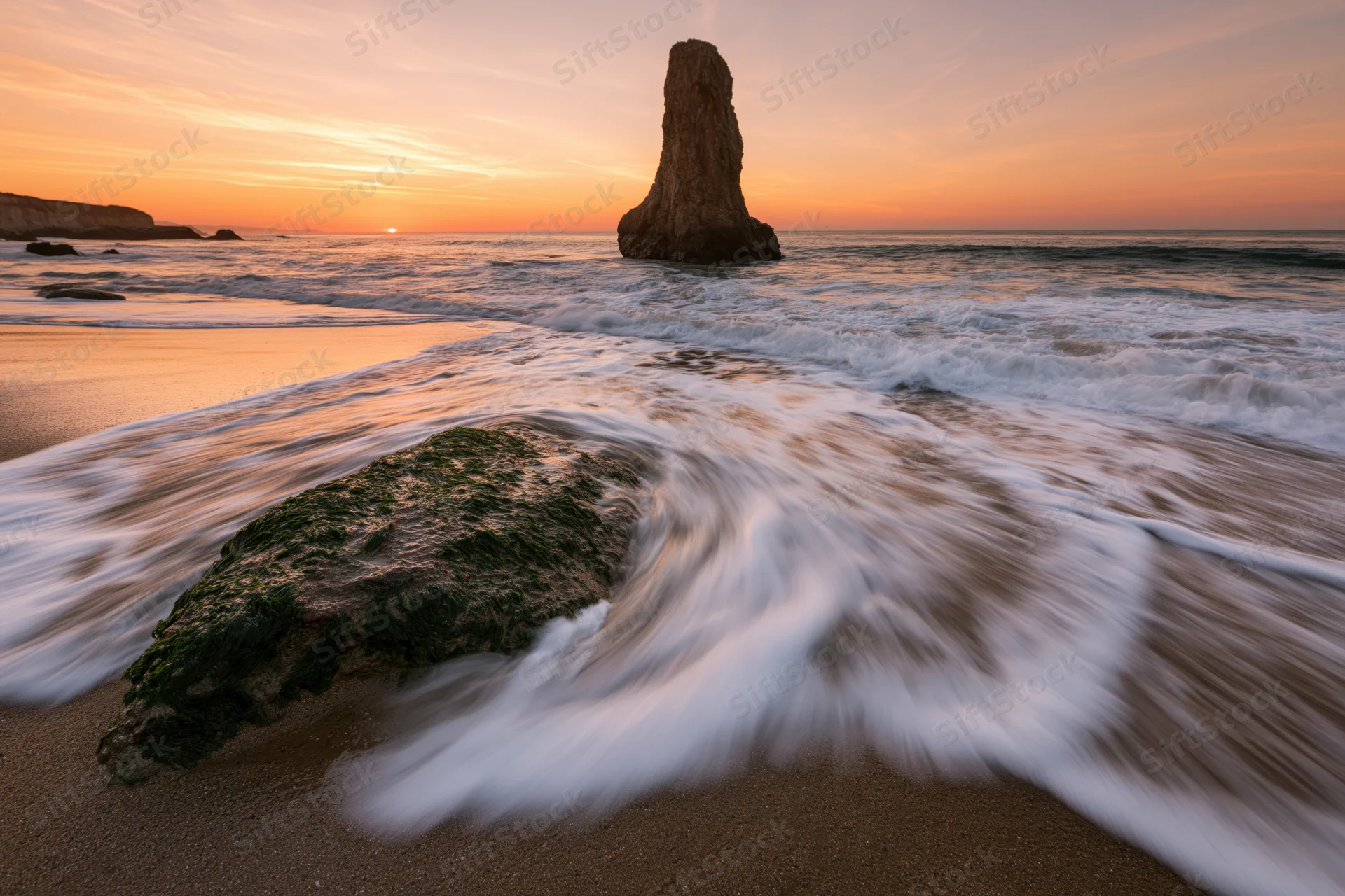 Ocean waves washing over moss-covered rock on sandy beach at sunrise
