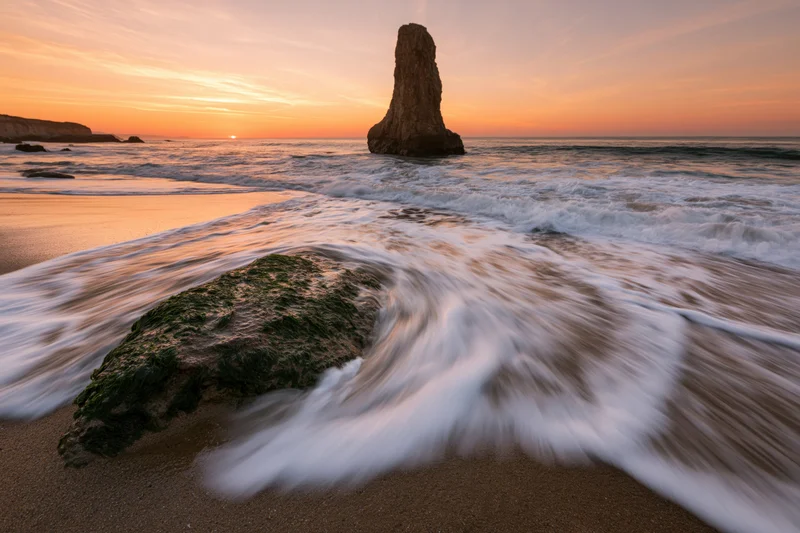 Ocean waves washing over moss-covered rock on sandy beach at sunrise