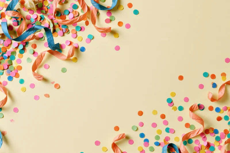Colorful confetti dots and curled paper streamers scattered across a pale yellow surface in a flat lay arrangement with copyspace
