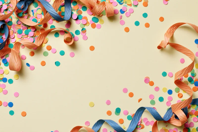 Colorful confetti dots and curled paper streamers scattered across a light beige surface forming a festive border with copyspace in the center