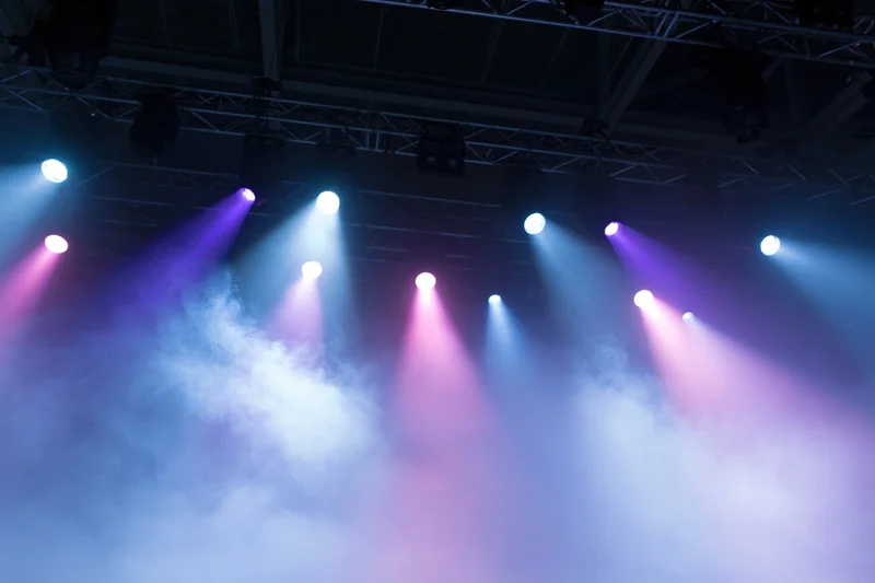 Stage lighting rig with multiple spotlights casting colored beams through smoke from below