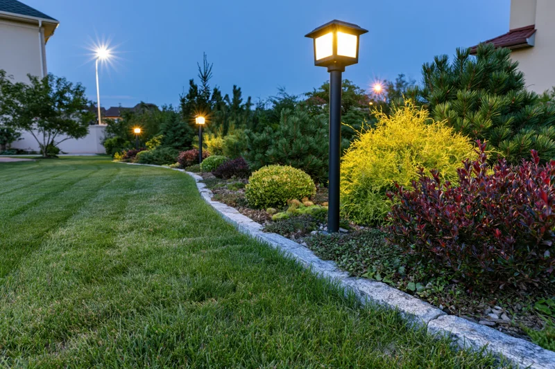 Curved stone-edged garden bed with mixed dwarf conifers and shrubs along a lawn pathway at twilight