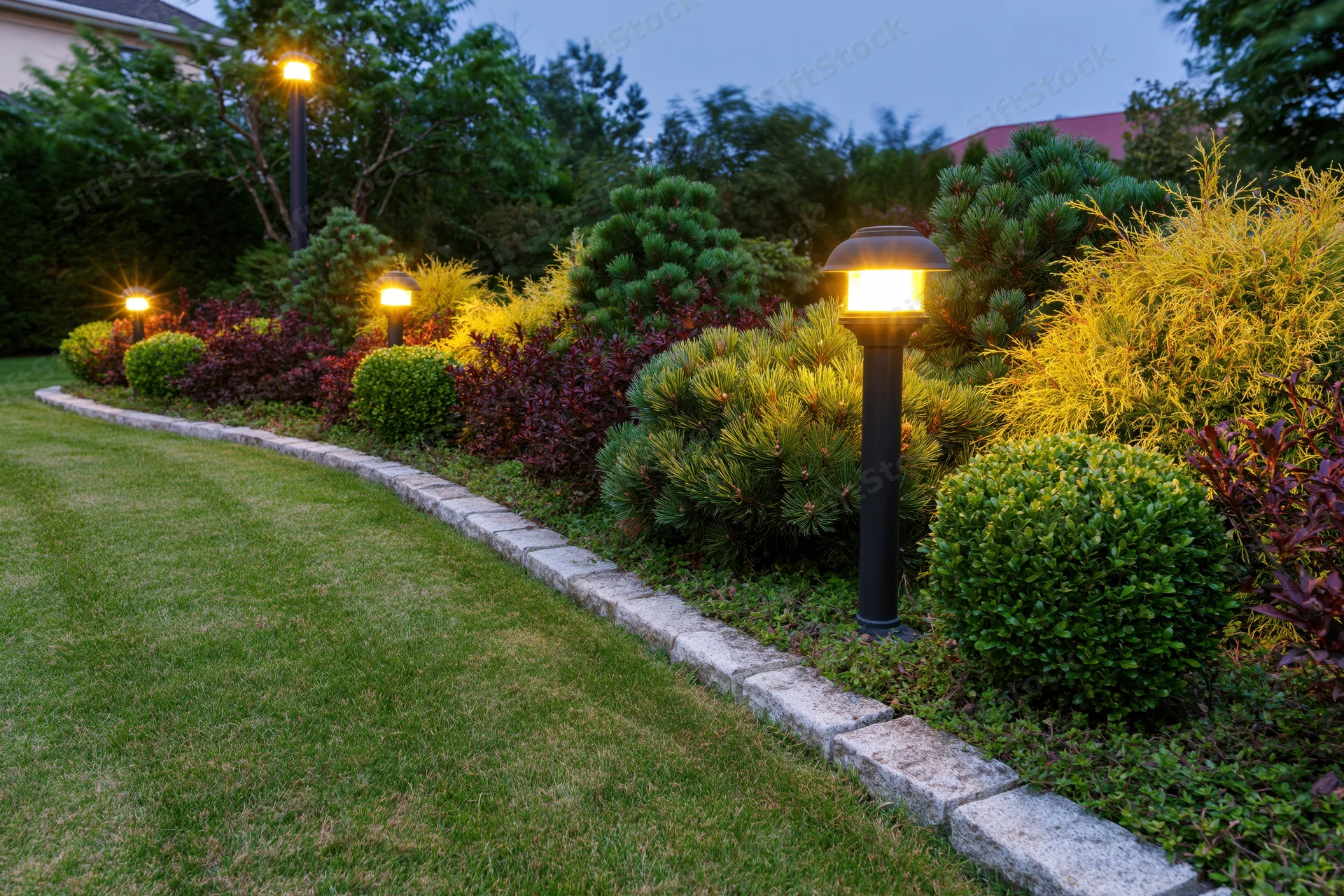 Curved stone-edged garden bed with mixed dwarf conifers and pathway lighting at dusk