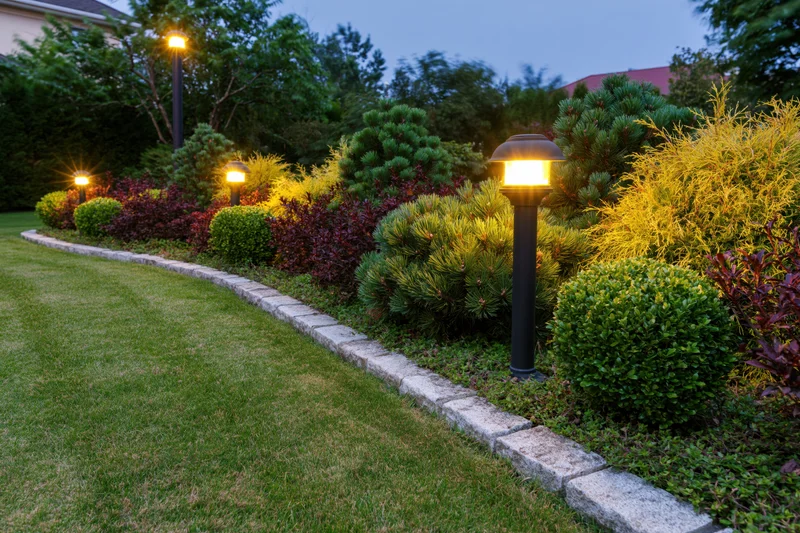 Curved stone-edged garden bed with mixed dwarf conifers and pathway lighting at dusk