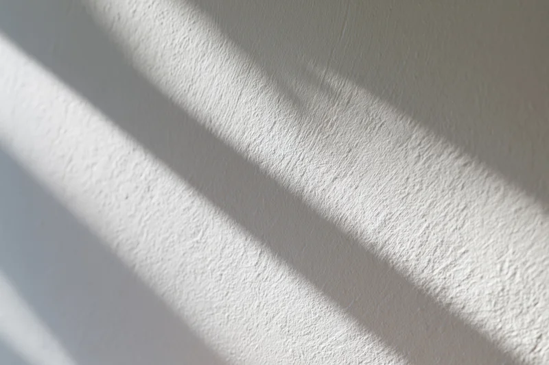 Diagonal shadows from natural light cast across a white textured wall in an interior space