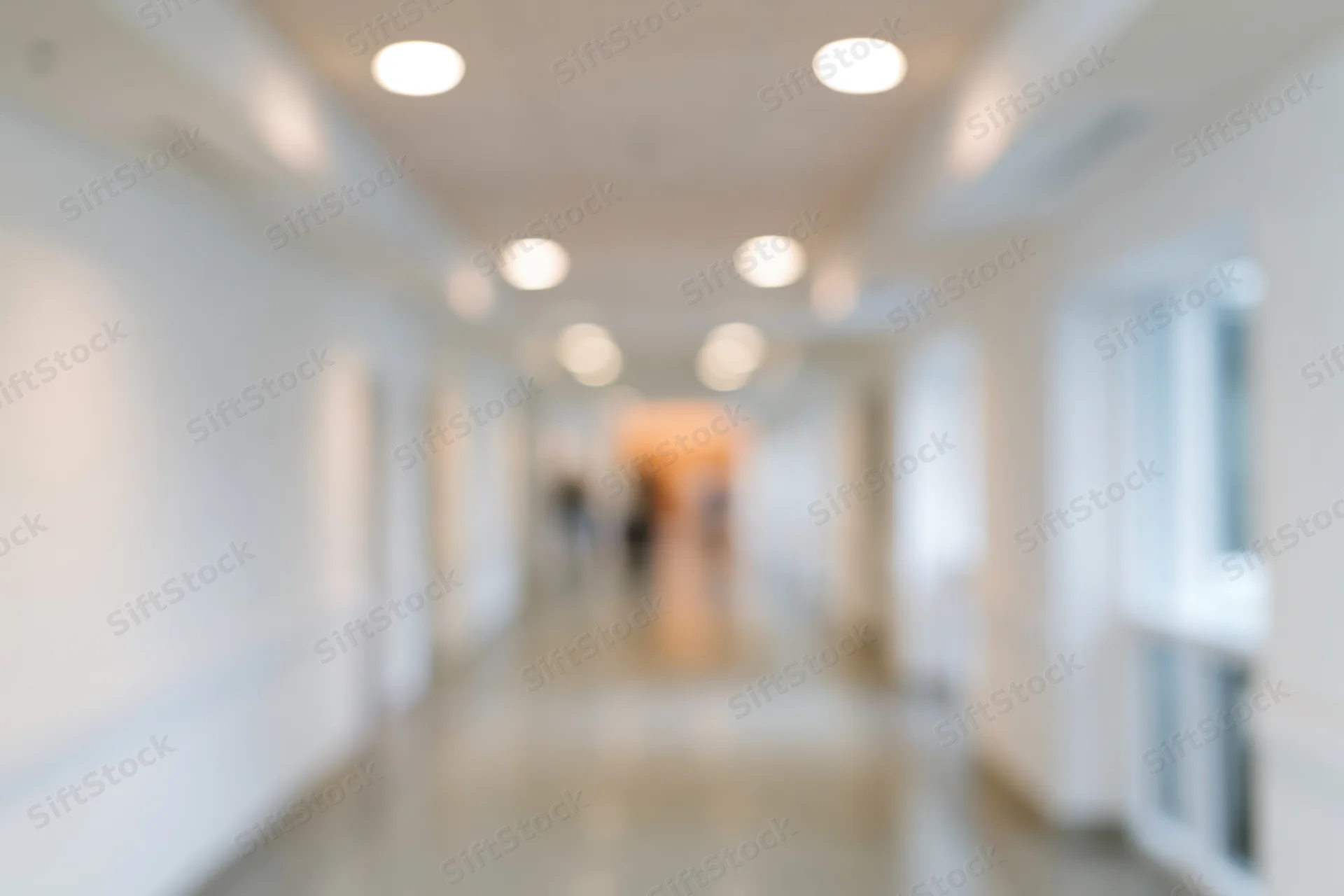 Defocused view of a long white hallway with recessed ceiling lights and reflective floor in a hospital or office building