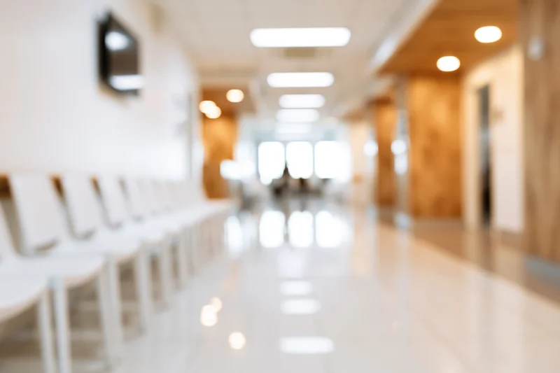 Defocused view of a hospital corridor with a row of white chairs along the wall