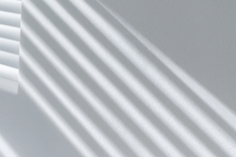 Diagonal shadows from window blinds cast across a white wall interior