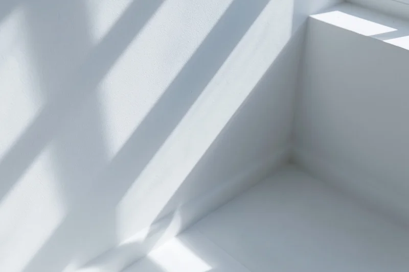 Diagonal window frame shadows and soft light patterns cast across white interior corner walls and floor
