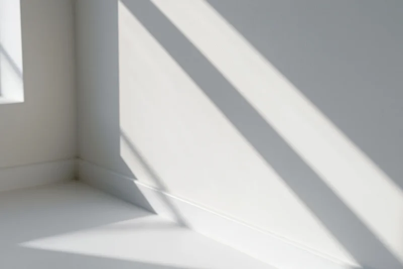 Diagonal shadows from a window cast across a white interior corner, creating a pattern of light and dark on walls and floor