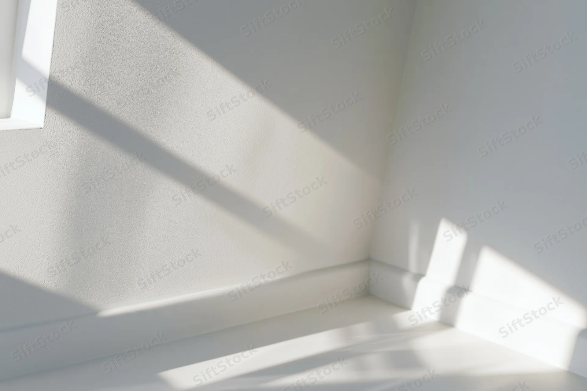 Diagonal window frame shadows and soft light patterns cast across white interior walls and floor in a corner of a minimalist room