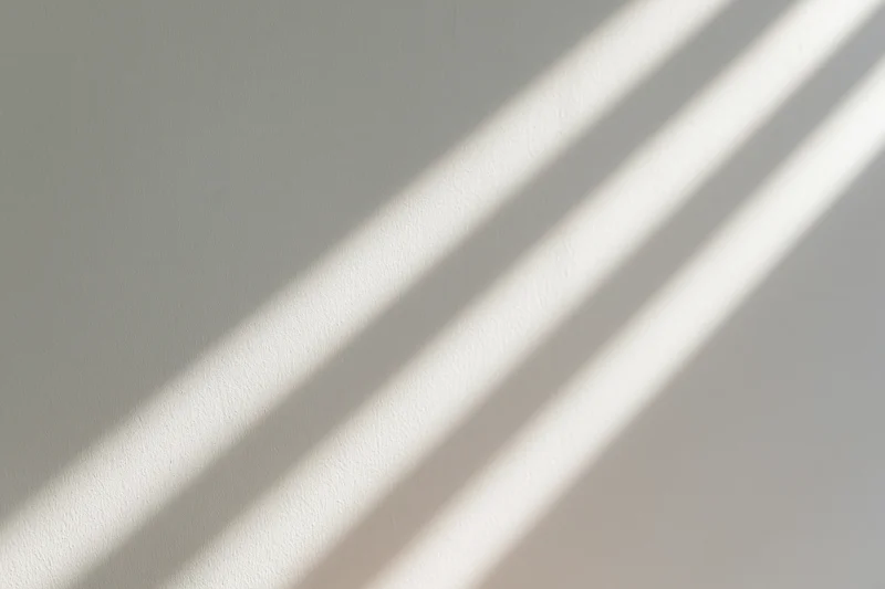 Diagonal stripes of sunlight and shadow cast across a plain white wall in an interior space