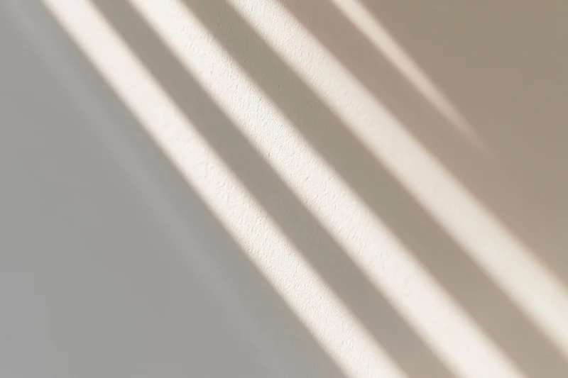 Diagonal stripes of sunlight and shadow cast across a plain wall indoors
