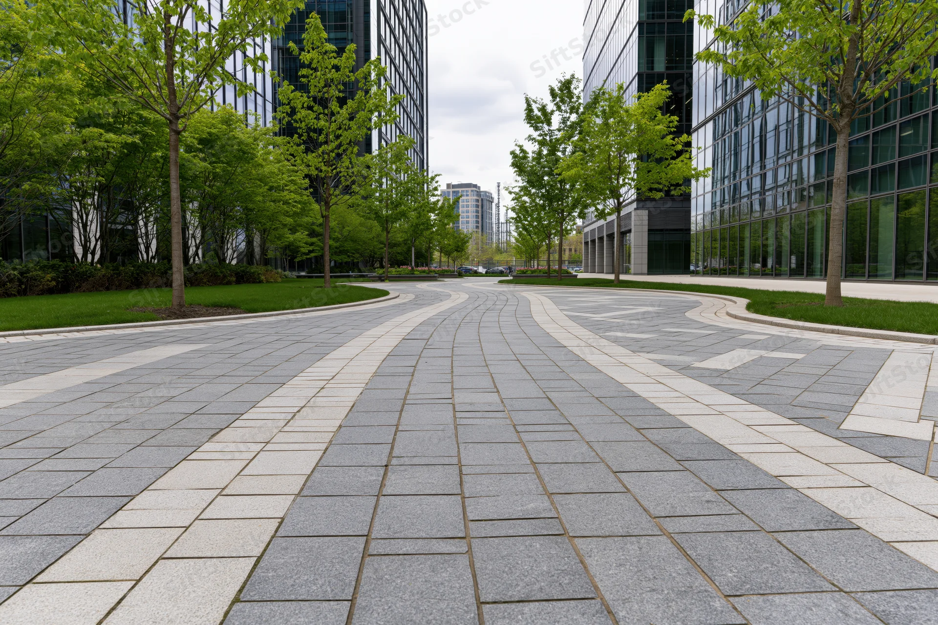 Curved stone-paved walkway winding between modern glass office buildings in an urban plaza with green trees and lawn