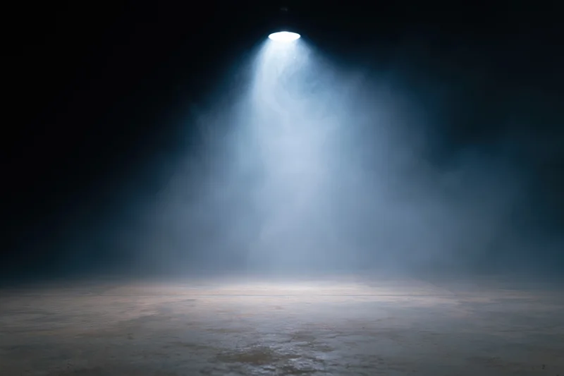 Single overhead spotlight illuminating an empty dark stage with fog and textured floor