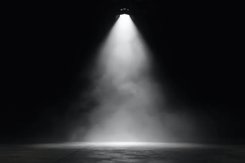 Single overhead spotlight illuminating empty dark stage with smoke and beam of light