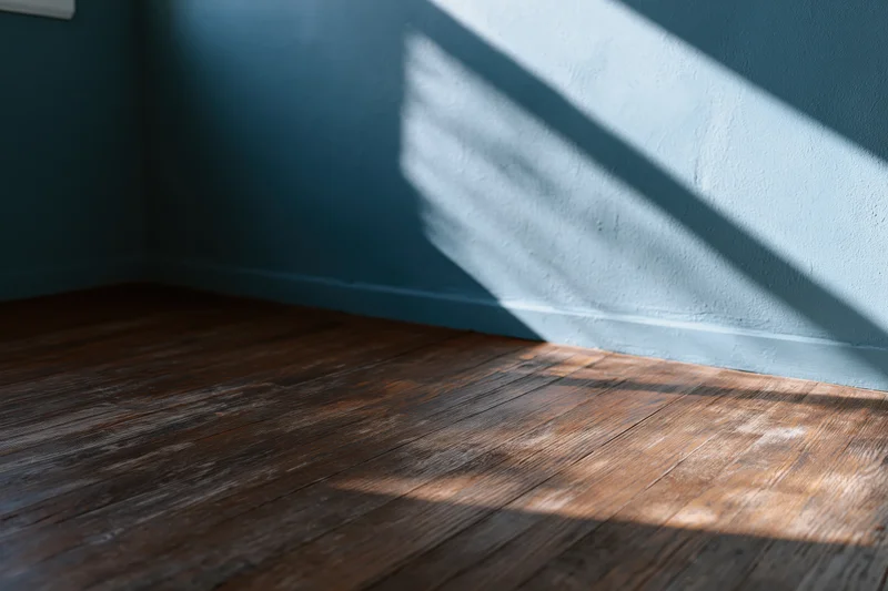Empty room corner with dark teal-grey painted wall and worn wooden floor lit by diagonal sunlight casting striped shadows
