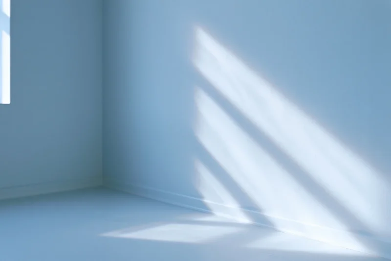 Empty minimalist room with pale blue-white walls and sunlight casting diagonal shadows in corner