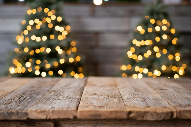 Rustic wooden tabletop in sharp focus with blurred christmas trees and warm bokeh lights in background, providing copyspace for product display