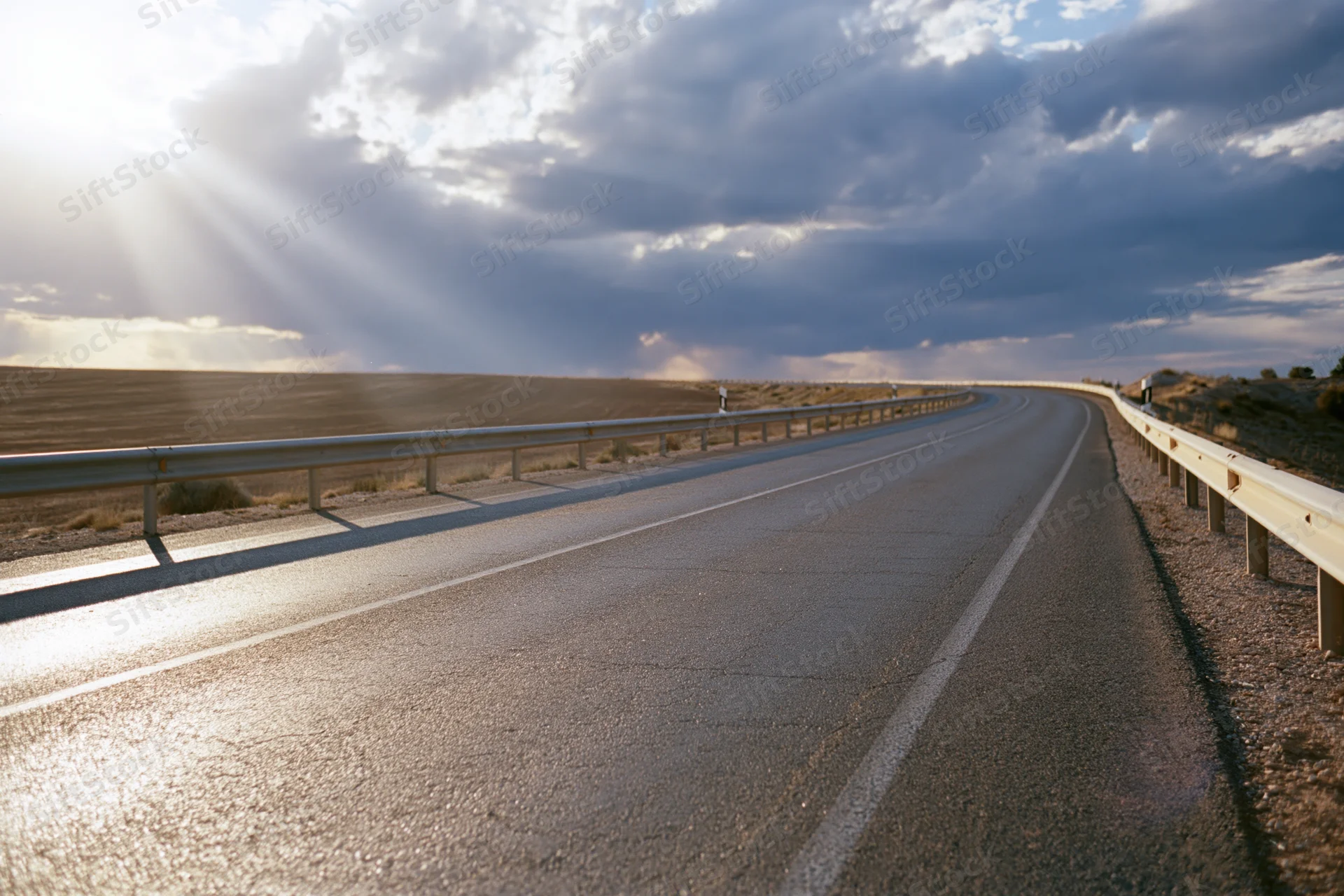Empty two-lane asphalt road curving gently through rural landscape under dramatic sky with sun rays breaking through clouds