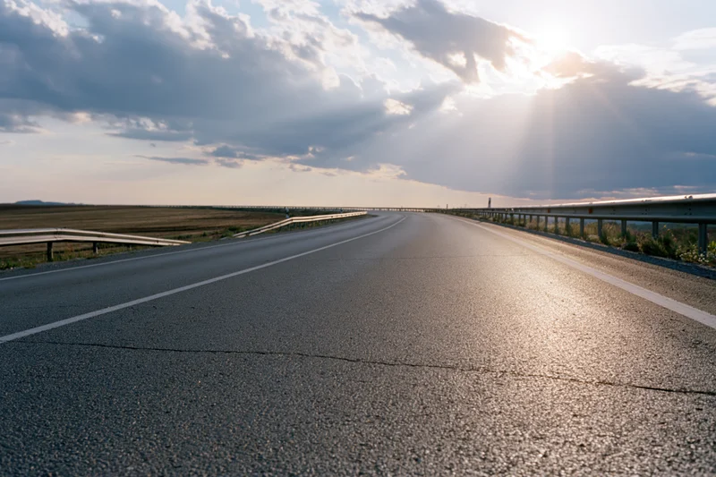 Empty two-lane asphalt road curving gently into the distance under a cloudy sky with sunbeams breaking through at sunset