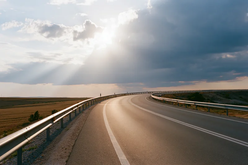Curving two-lane asphalt road through rural farmland under cloudy sky with sunbeams breaking through