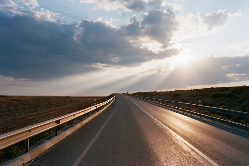 Empty two-lane asphalt road curving into the distance under a dramatic sunset sky with sunbeams breaking through clouds