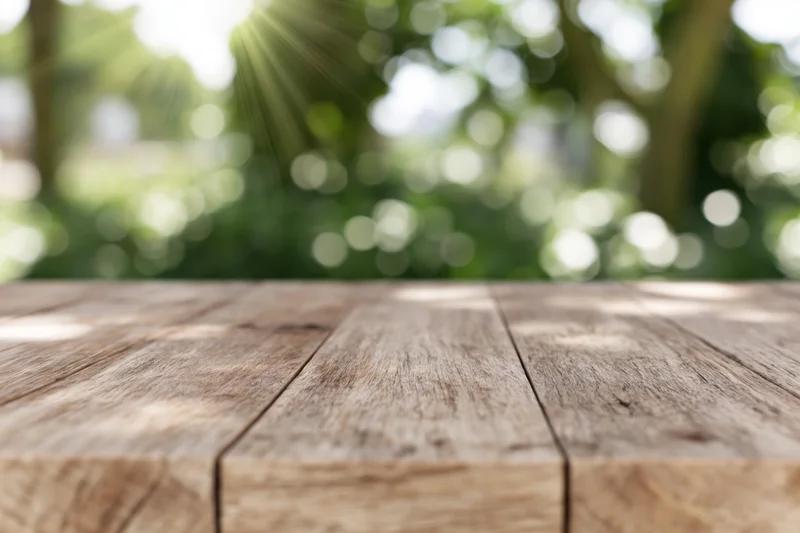 Weathered wooden tabletop in outdoor setting with blurred green foliage and sunlight in background
