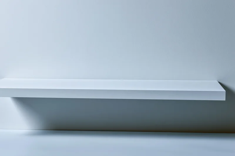 Empty white shelf mounted on a plain light gray wall in a studio setting