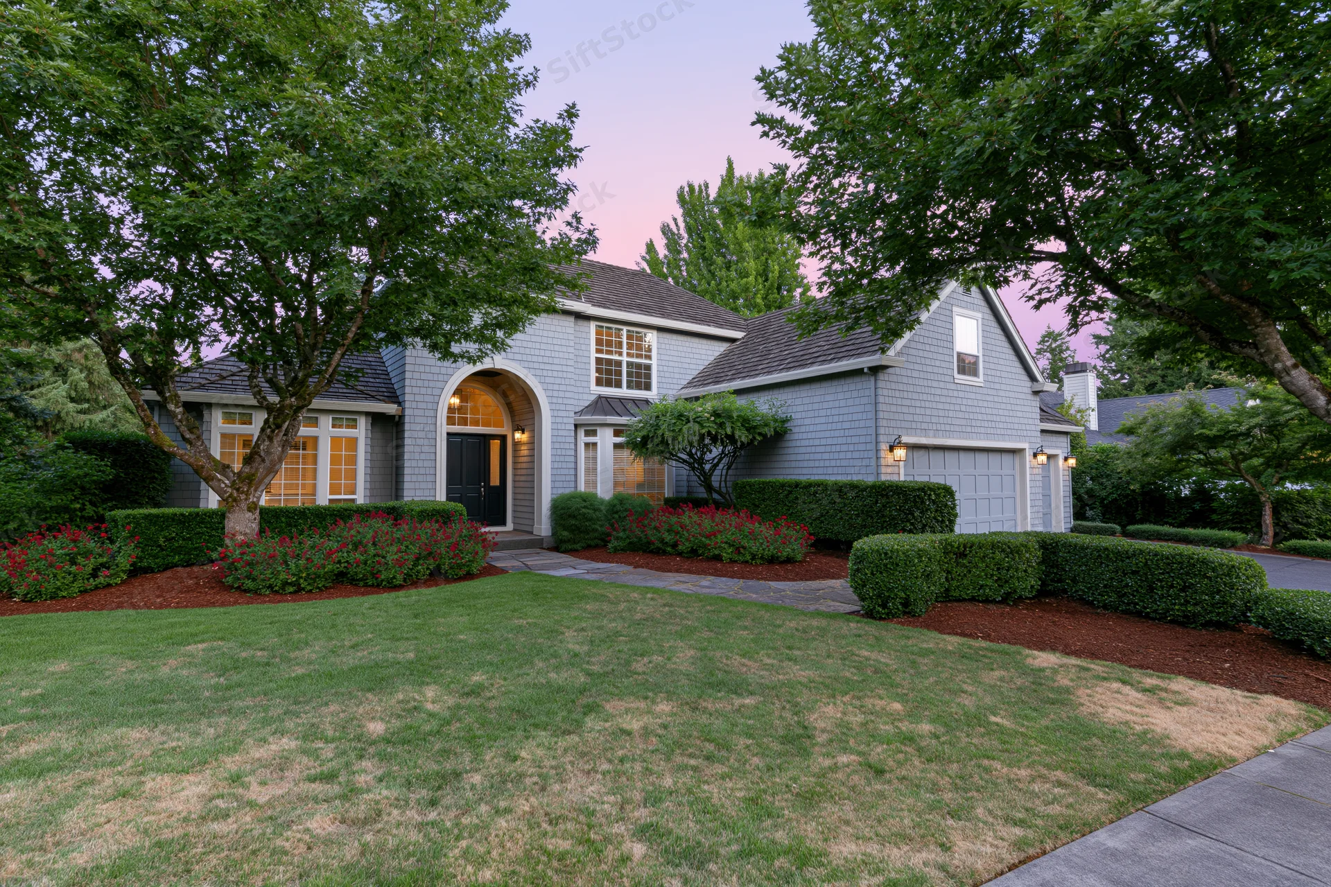 Gray cedar-sided suburban house with arched entrance and landscaped front yard at dusk