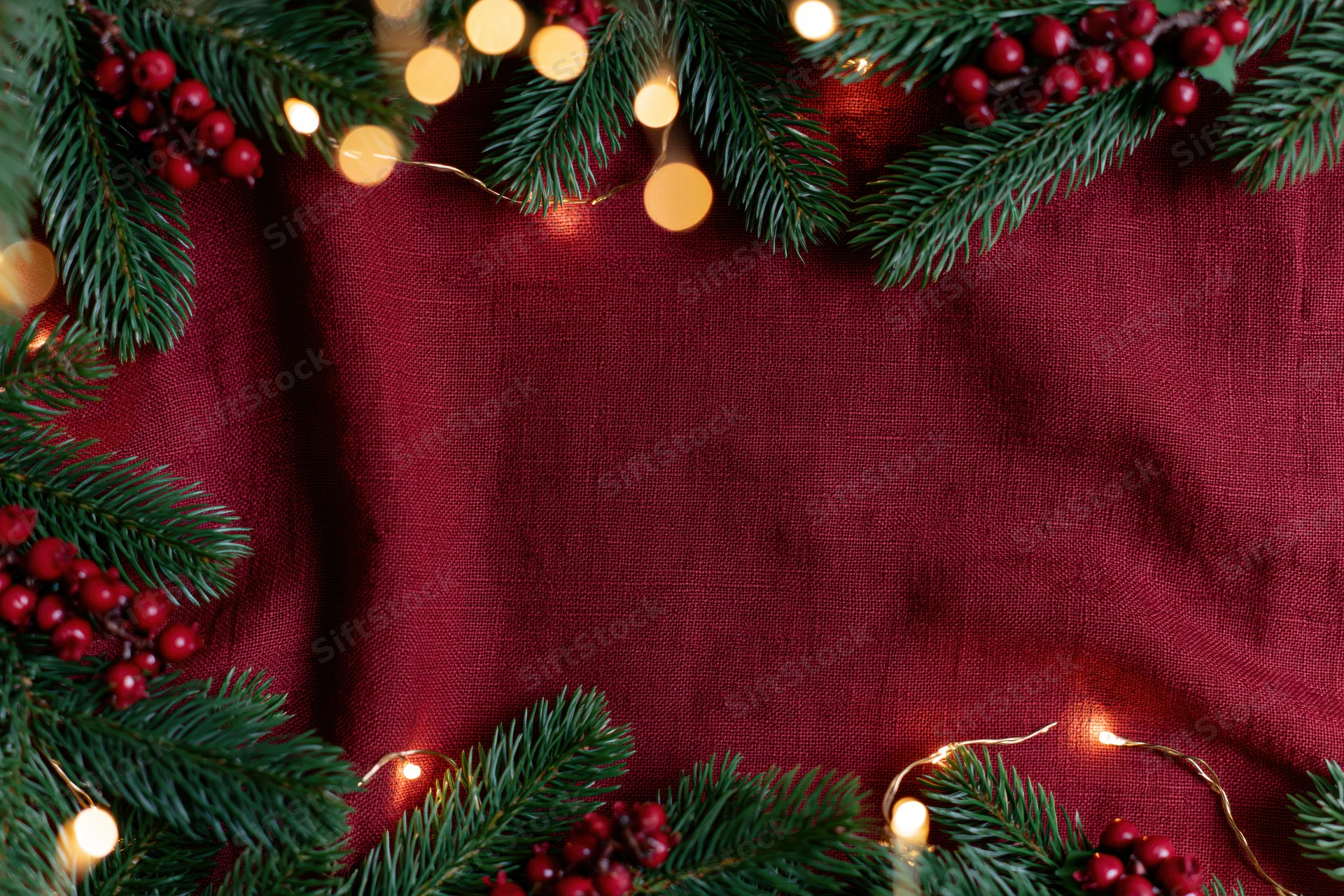 Pine branches and holly berries arranged in a flat lay with string lights on a red fabric background