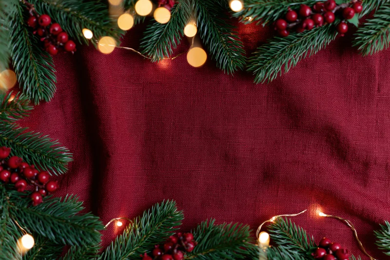 Pine branches and holly berries arranged in a flat lay with string lights on a red fabric background