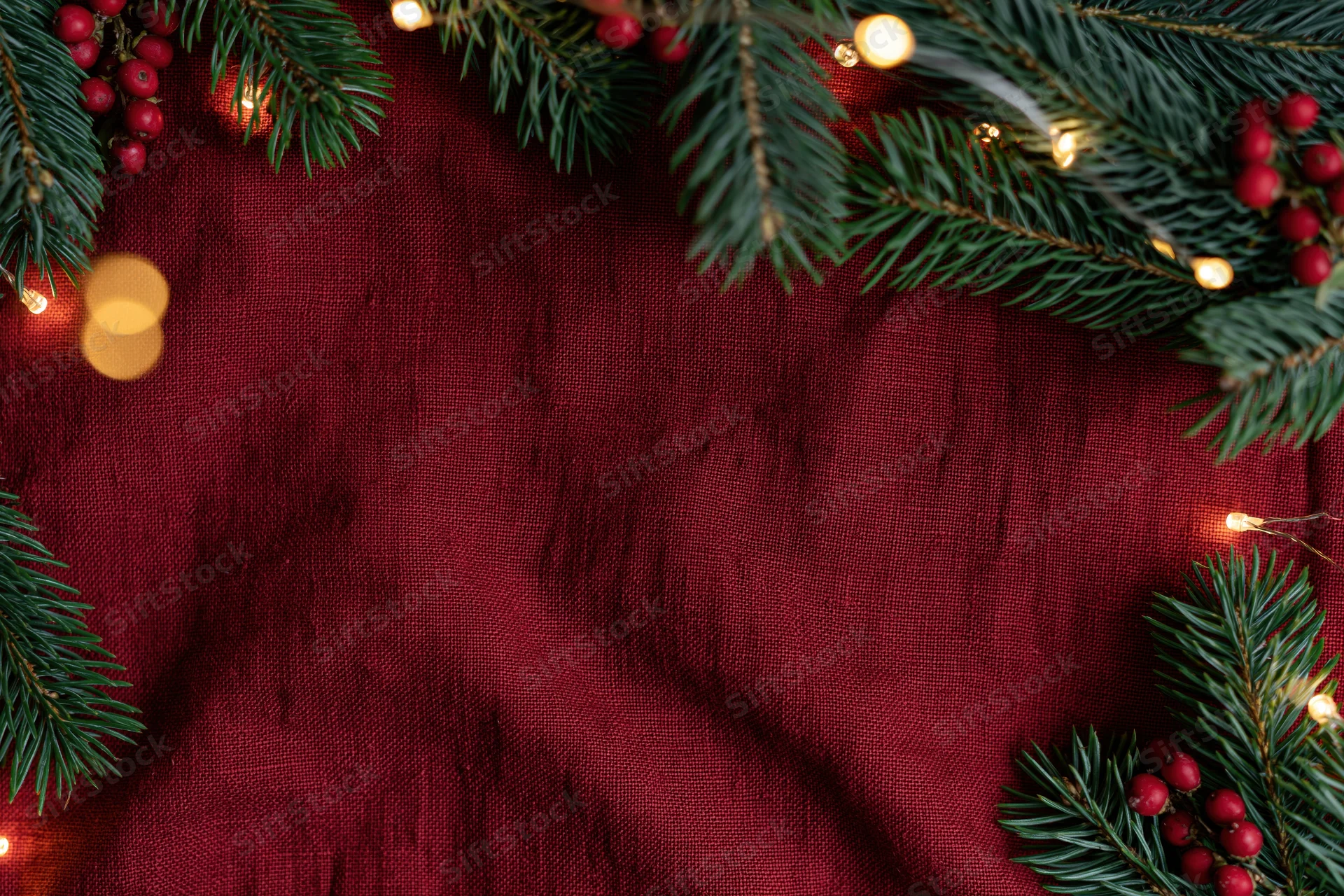 Pine branches and holly berries arranged in a flat lay on red fabric with string lights forming a border