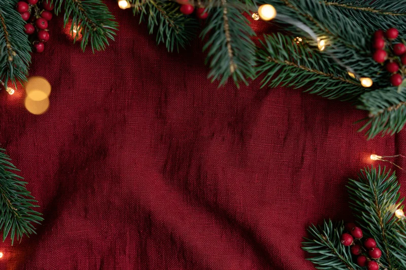 Pine branches and holly berries arranged in a flat lay on red fabric with string lights forming a border
