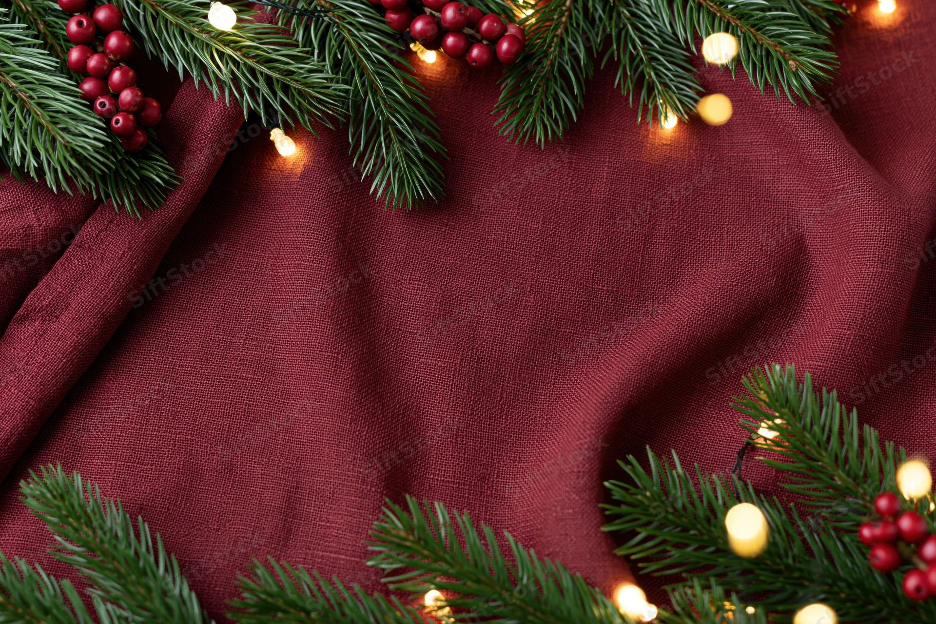 Pine branches and holly berries arranged with string lights on maroon fabric in flat lay composition