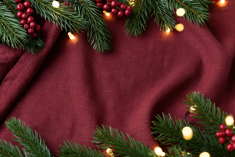 Pine branches and holly berries arranged with string lights on maroon fabric in flat lay composition