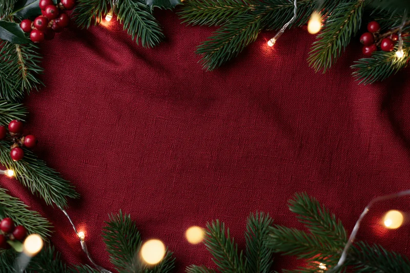 Pine branches and holly berries arranged in a frame with string lights on a red fabric background