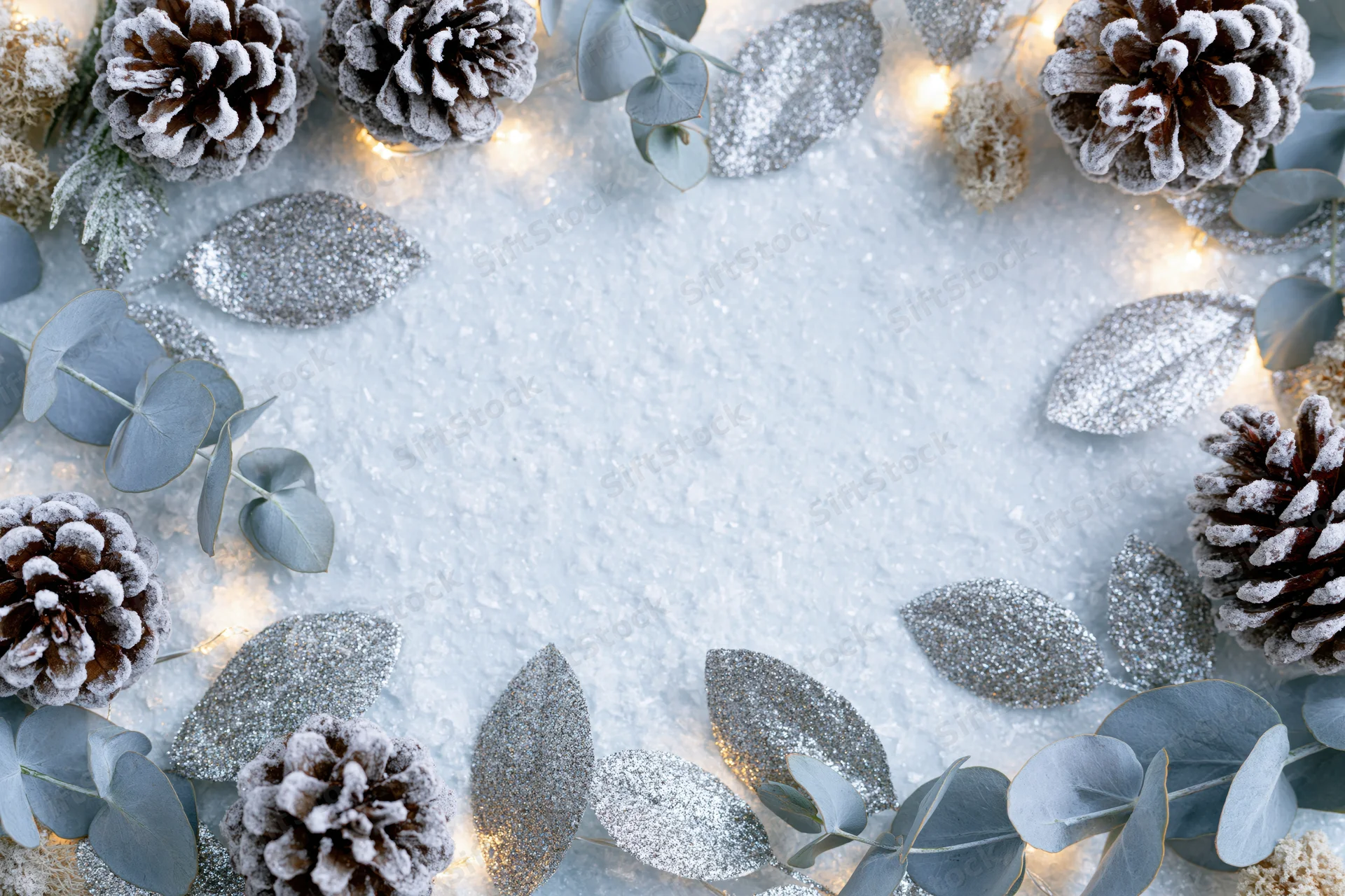 Frosted pinecones and eucalyptus sprigs arranged in a flat lay frame with silver glitter leaves and warm lights on a snowy surface