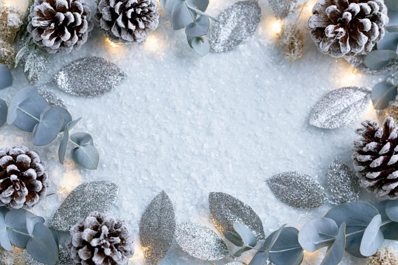 Frosted pinecones and eucalyptus sprigs arranged in a flat lay frame with silver glitter leaves and warm lights on a snowy surface