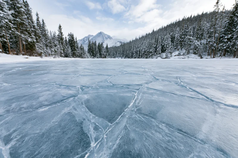 Cracked blue ice covering a frozen alpine lake surrounded by snow-covered pine trees and distant mountains