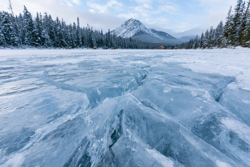 Cracked blue ice on frozen alpine lake surrounded by snow-covered pine forest and distant mountain peak