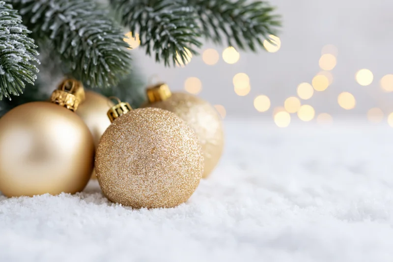 Gold christmas ornaments resting in soft white snow beneath frosted pine branches with blurred holiday lights in background