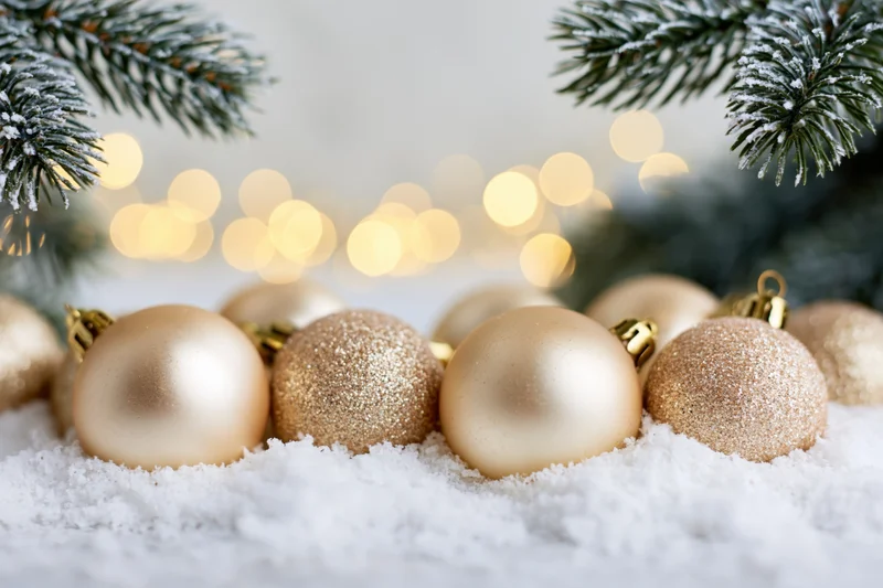 Gold christmas ornaments resting in soft white snow beneath frosted pine branches with blurred warm lights in background