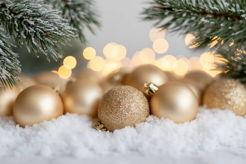 Gold christmas ornaments resting in soft white snow beneath frost-covered pine branches with warm bokeh lights in background